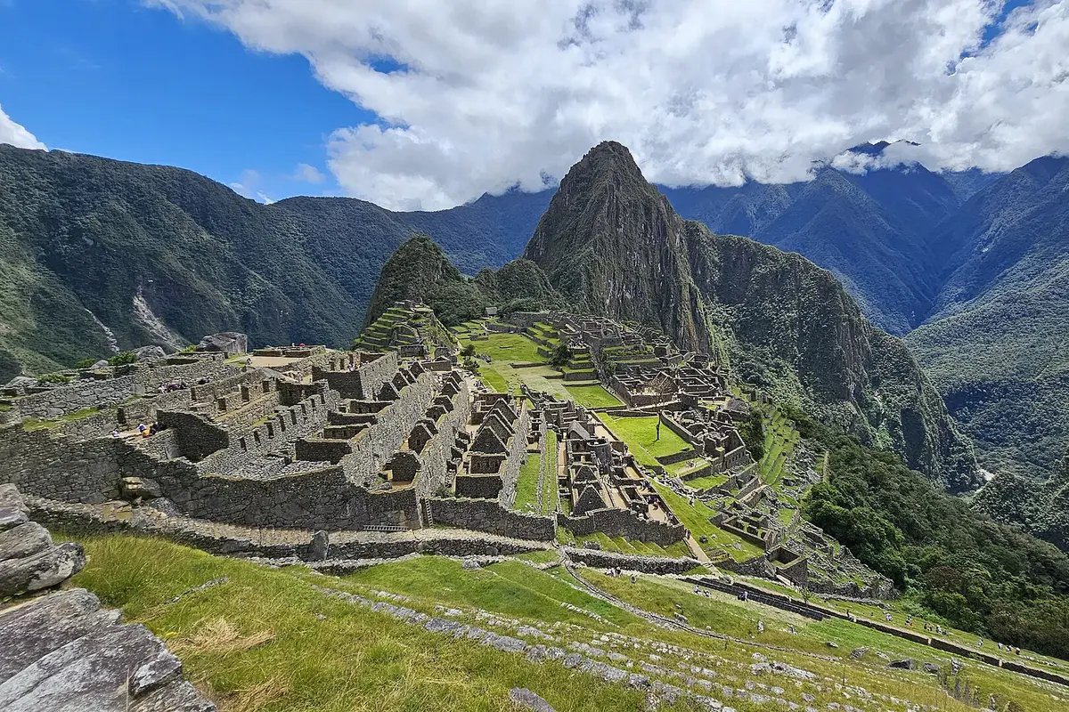 Le terrazze in pietra e gli edifici di Machu Picchu su una cresta verdeggiante, con la ripida vetta di Huayna Picchu che si erge dietro sotto nuvole sparse