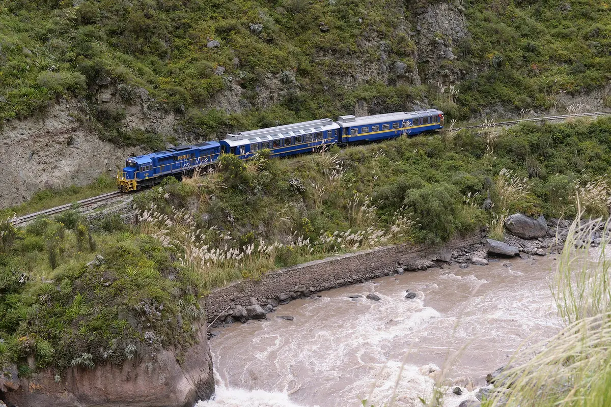 Ein blau-gelber PeruRail-Zug, der sich an einem grünen Hang über dem braunen, schnell fließenden Urubamba-Fluss entlangschlängelt